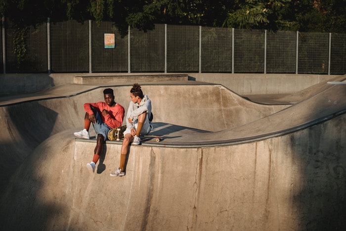 Two people sitting on the edge of a skate park with a skateboard.